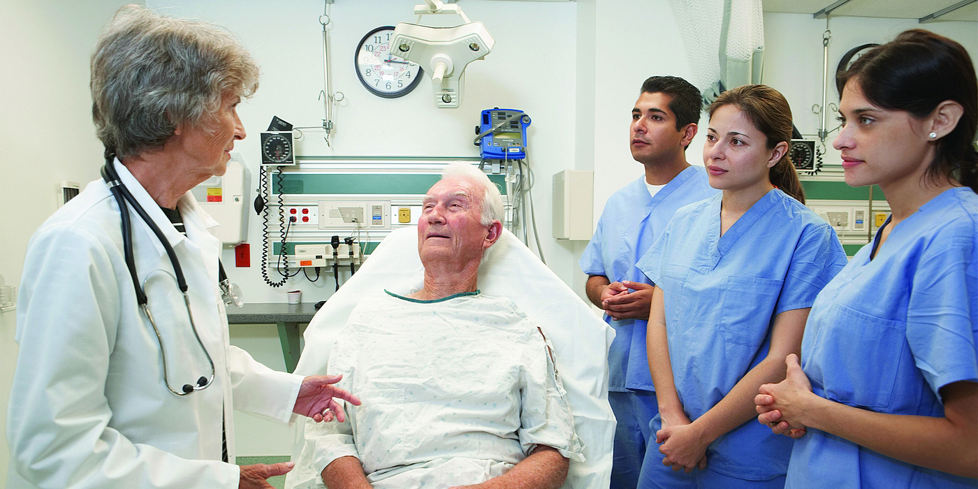 A physician discusses a patient’s case with medical students, while patient sits up in a hospital bed.